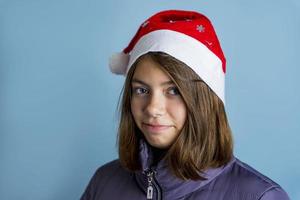 A beautiful girl in a Christmas cap and a warm jacket on a blue background looks at the camera photo