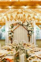 Festive setting table with meals for wedding, wooden arch, stands decorated with composition of white flowers and greenery in the rustic banquet hall. photo