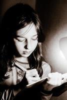 A teenage girl writes in a notebook with a pen. Black and white photo of a girl lit by a table lamp.