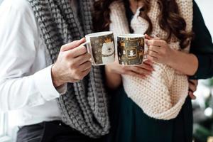 Christmas. Love. Home. Young couple in warm clothes is holding in hands cups with hot coffee or tea at home near the Christmas tree photo