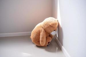 The teddy bear is wearing a mask, sitting and bowed down in the corner of the room. Depression, sad, disappointment or illness photo