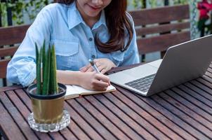 A beautiful woman with long hair sitting and writing notes in a book on a desk. Woman working or studying via using a notebook computer. Relaxing working or studying online at home. photo