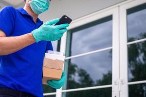 The delivery man holding a paper box, wears a mask and wears rubber gloves stand front home of customer and check order online in smartphone. Transportation Fast and free delivery online shopping. photo