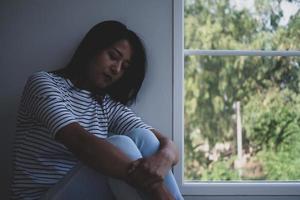 An Asian woman sat alone in the house with her knees beside the window. With sad and disappointed faces and psychological symptoms. Depression or an unexpected pregnancy concept. photo