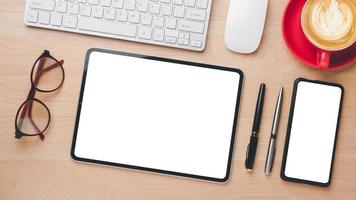 Office wooden desk workplace with blank sceen tablet and smartphone, keyboard, mouse, eyeglass, pen and cup of coffee, Top view flat lay. photo