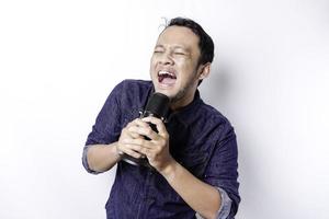 Portrait of carefree Asian man, having fun karaoke, singing in microphone while standing over white background photo