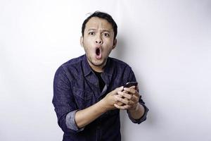 hombre asiático conmocionado con camisa azul y sosteniendo su teléfono, aislado de fondo blanco foto