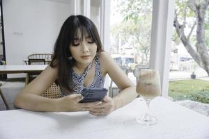 Sad face of a young Asian woman holding a cell phone wearing a tank top sitting near a window. Advertisement concept. photo