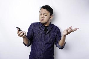 A thoughtful young Asian man is wearing blue shirt holding his phone and looks confused, isolated by white background photo