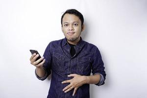 A thoughtful Asian man is hungry and touching his belly while holding his phone thinking what to order, isolated by white background photo