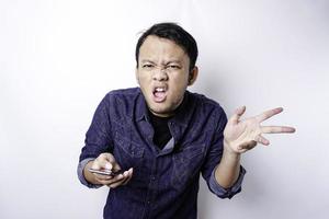 The angry and mad face of Asian man in blue shirt holding his phone isolated white background.
