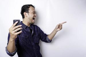 Excited Asian man wearing blue shirt pointing at the copy space beside him while holding his phone, isolated by white background photo