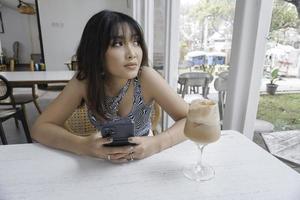Sad face of a young Asian woman holding a cell phone wearing a tank top sitting near a window. Advertisement concept. photo