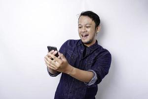 Excited Asian man wearing blue shirt smiling while holding his phone, isolated by white background photo