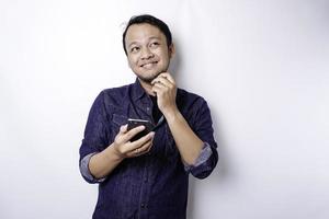 A thoughtful young man dressed in blue shirt while looking aside holding his phone, isolated by white background photo