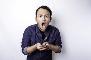 hombre asiático conmocionado con camisa azul y sosteniendo su teléfono, aislado de fondo blanco foto