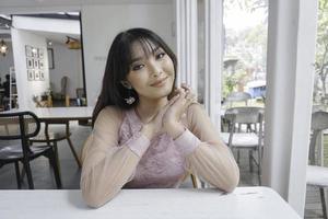 A smiling Asian woman wearing pink dress while sitting near window photo