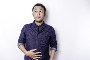 An Asian man is hungry and touching his belly while looking aside thinking what to eat, isolated by white background photo