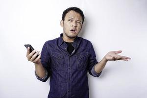 un joven asiático pensativo lleva un pantalones azul sosteniendo su teléfono y parece confundido, aislado por un fondo blanco foto