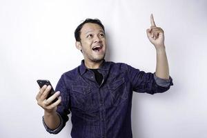 un retrato de un hombre asiático feliz con una camisa azul y sosteniendo su teléfono, aislado por un fondo blanco foto