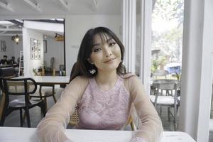 A smiling Asian woman wearing pink dress while sitting near window photo