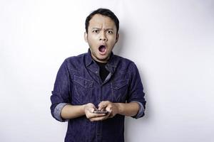 Shocked Asian man wearing blue shirt and holding his phone, isolated by white background photo