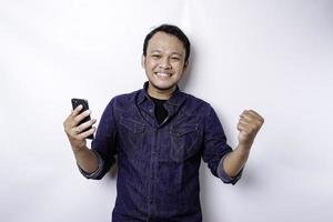 un joven asiático con una expresión feliz y exitosa que usa pantalones azul y sostiene su teléfono, aislado de fondo blanco foto
