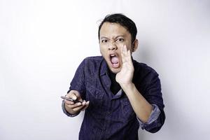 The angry and mad face of Asian man in blue shirt holding his phone isolated white background. photo