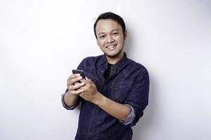 Excited Asian man wearing blue shirt smiling while holding his phone, isolated by white background photo