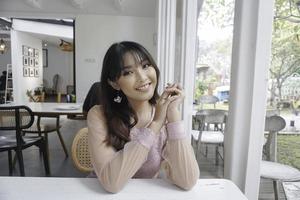 A smiling Asian woman wearing pink dress while sitting near window photo