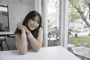 A smiling Asian woman wearing pink dress while sitting near window photo