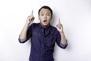 Shocked Asian man wearing blue shirt pointing at the copy space on top of him, isolated by white background photo