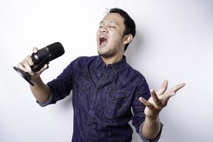 Portrait of carefree Asian man, having fun karaoke, singing in microphone while standing over white background photo