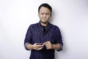 A thoughtful young Asian man is wearing blue shirt holding his phone and looks confused, isolated by white background photo