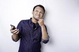 A thoughtful young man dressed in blue shirt while looking aside holding his phone, isolated by white background photo