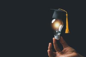 academy and success graduate education concept. businessman hand holding bright, electric light bulb with degree cap on black background. business education, knowledge, learning idea with copy space. photo