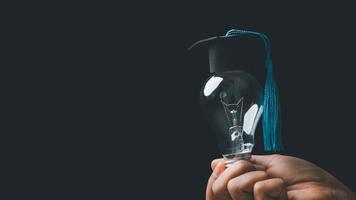 academy and success graduate education concept. businessman hand holding bright, electric light bulb with degree cap on black background. business education, knowledge, learning idea with copy space. photo