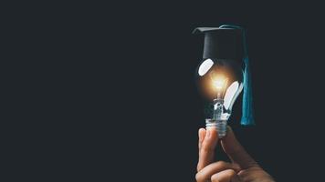 academy and success graduate education concept. businessman hand holding bright, electric light bulb with degree cap on black background. business education, knowledge, learning idea with copy space. photo