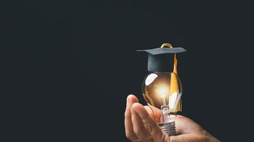 academy and success graduate education concept. businessman hand holding bright, electric light bulb with degree cap on black background. business education, knowledge, learning idea with copy space. photo