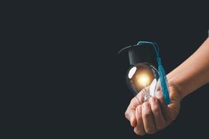 academy and success graduate education concept. businessman hand holding bright, electric light bulb with degree cap on black background. business education, knowledge, learning idea with copy space. photo