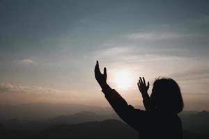 cristianismo de fe del concepto de adoración cristiana. la oración espiritual entrega el brillo del sol con un hermoso fondo de puesta de sol borroso. mujer rezando a dios con bendición esperanzadora contra la puesta de sol. foto