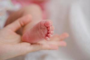 Close up mother holding foot newborn girl in a room. Adorable infant rests on white bedsheets, staring at camera looking peaceful. Infancy, healthcare and paediatrics, babyhood concept. photo