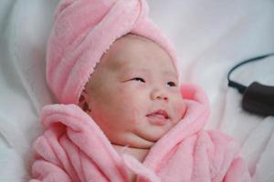 Closeup cute newborn baby in pink bodysuit lying down alone on bed. Adorable infant rests on white bedsheets, staring at camera looking peaceful. Infancy, healthcare and paediatrics, babyhood concept photo