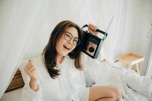 Funny young asian woman with beaming smile in eyewear holding retro style radio on shoulder. listening to music singing and dancing on bed at home. She wears white shirt. Music lover fan hobby concept photo