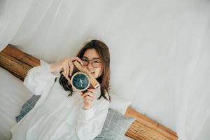 retrato de mujer hermosa asiática feliz sonriendo en camisa blanca posando en la cama. tomando fotos de un reloj que parece una cámara de fotos. concepto de estilo de vida de las personas. copie el espacio mirando a la cámara.