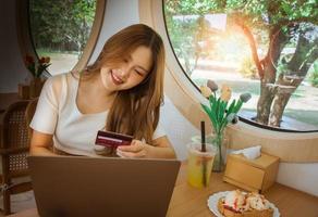 Young Asian woman holding credit card and using laptop computer. Beautiful happy young girl in cafe. Online shopping, E-commerce, spending money, technology money wallet and online payment concept.