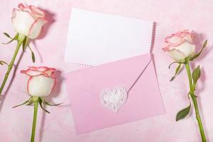 Pink envelope with empty white blank sheet and pale pink roses on pink backdrop. Copy space Top view photo