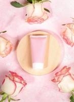 Pink blank cosmetic tube on wooden slice inside frame of pale pink roses on pink Top view Copy space photo