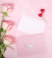 Festive flat lay arrangement of pale pink roses, envelope, hearts, empty white blank sheet on pink. photo