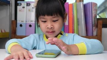 linda niña sosteniendo un teléfono inteligente mirando la pantalla sonriendo usando la aplicación, jugando juegos en línea, viendo dibujos animados en la sala de estar en casa. los niños y el concepto de tecnología moderna video